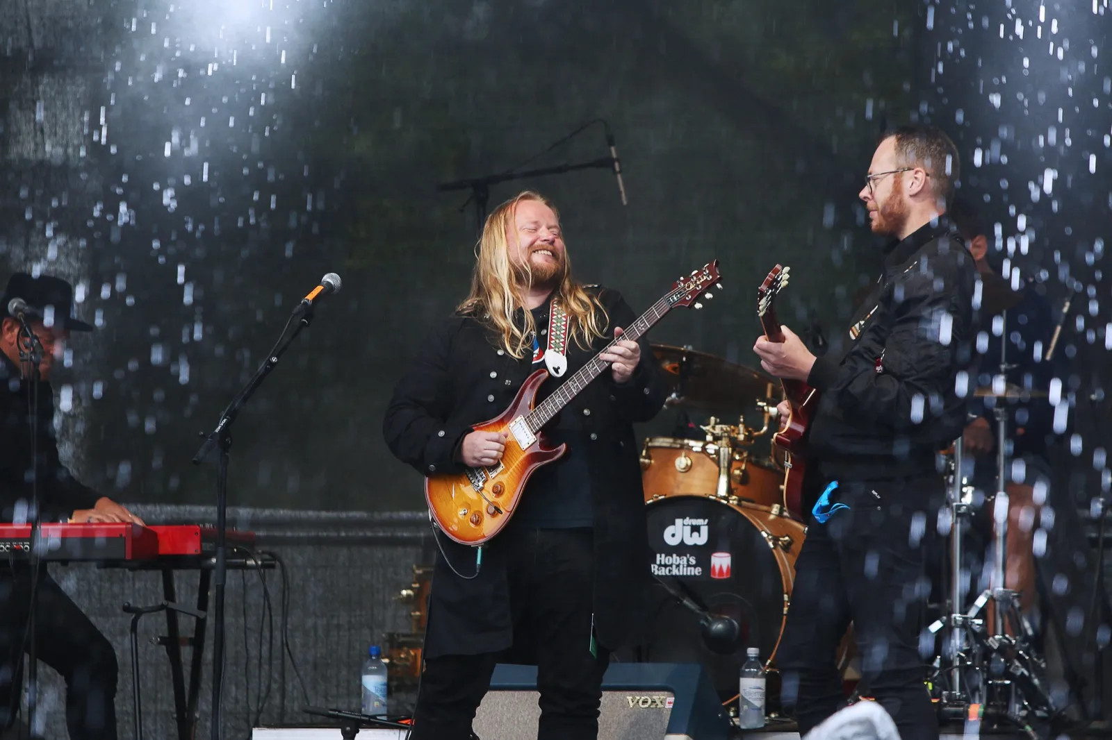 Nerli There på scenen i regnvær. Mats Nerli spiller gitar midt i bildet og ler, med gitarist til høyre og tangenter til venstre. Regndråpene lyser opp i motlyset.