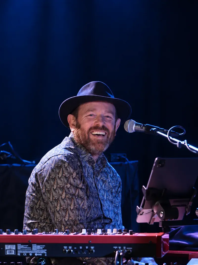 André Gussiås ved tangentene med hatt, smilende mot mikrofonen